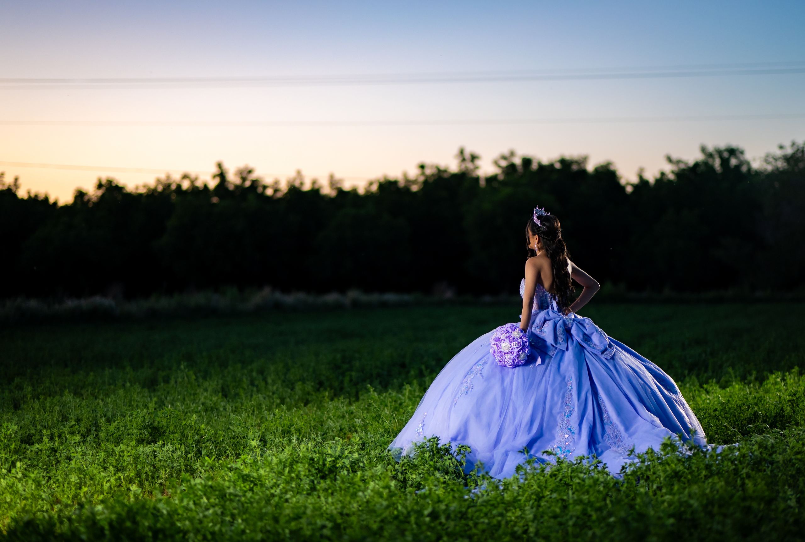 Quinceañera in a lavender gown standing in a green field at sunset